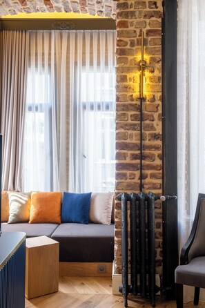 A room with a brick wall, a radiator, a sofa with colorful cushions, a wooden side table, and a chair. - Balo (Istanbul)