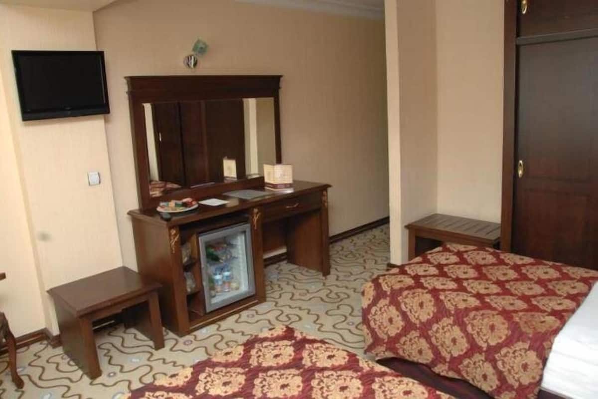 A hotel room with a wooden desk, a mirror, a television mounted on the wall, a small refrigerator, a patterned bedspread, and a door.