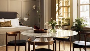 Une chambre d’hôtel avec une table de salle à manger ronde, quatre chaises, un lit avec une tête de lit, un luminaire suspendu et une fenêtre avec des rideaux.