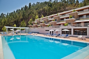 A swimming pool with sun loungers and umbrellas, surrounded by a building with multiple balconies and a forested hillside in the background.