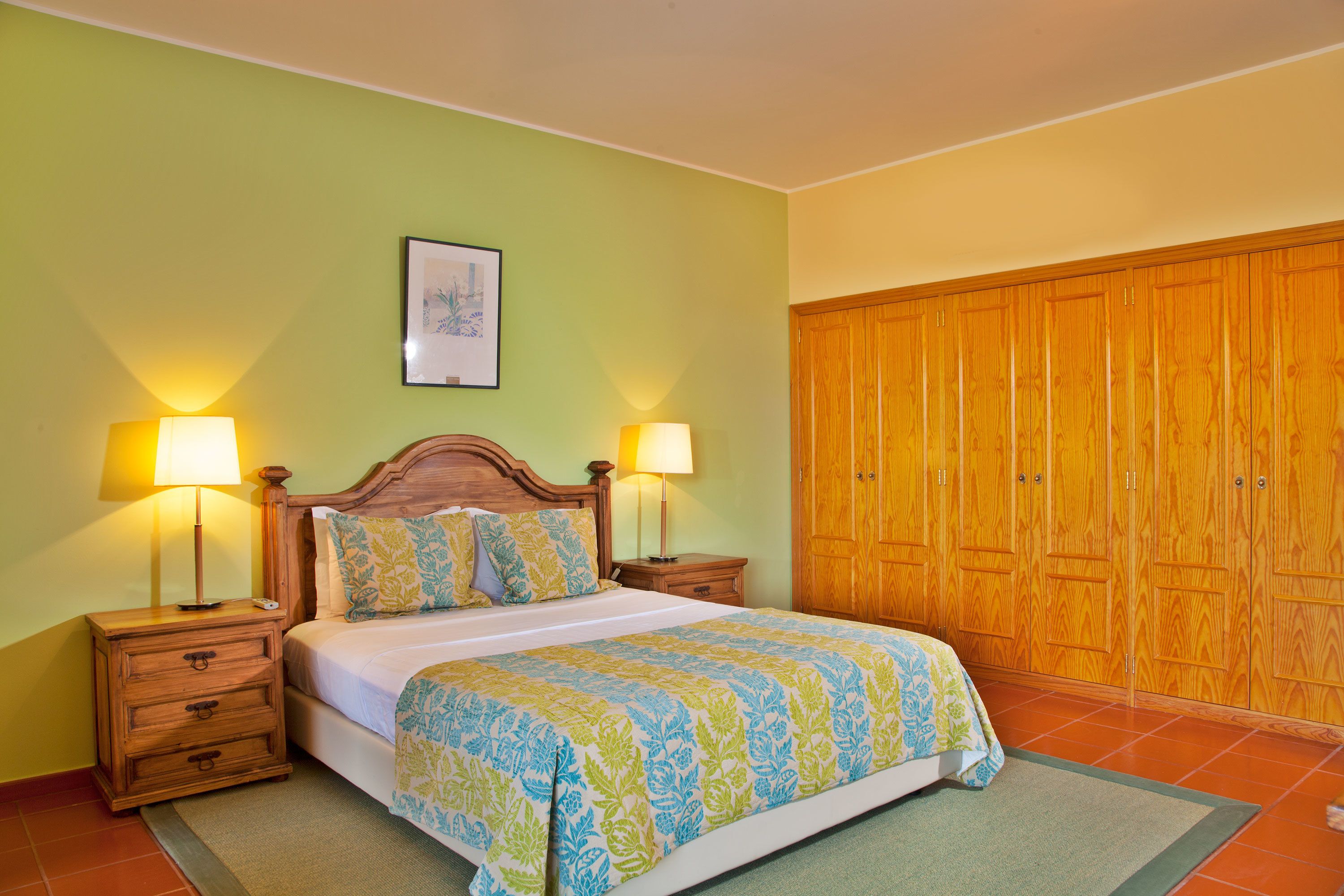 A bedroom with a bed, two bedside tables with lamps, a wooden wardrobe, and a framed picture on the wall.