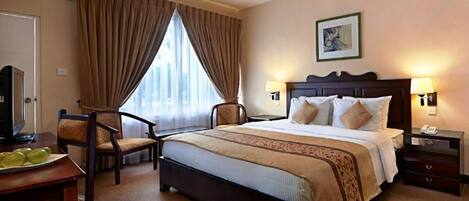 A hotel room with a large bed, two bedside tables with lamps, a desk with a chair, a television, and a window with curtains.