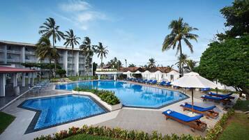 Ein Swimmingpool mit Liegestühlen und Sonnenschirmen, umgeben von Palmen und einem Hotelgebäude.
