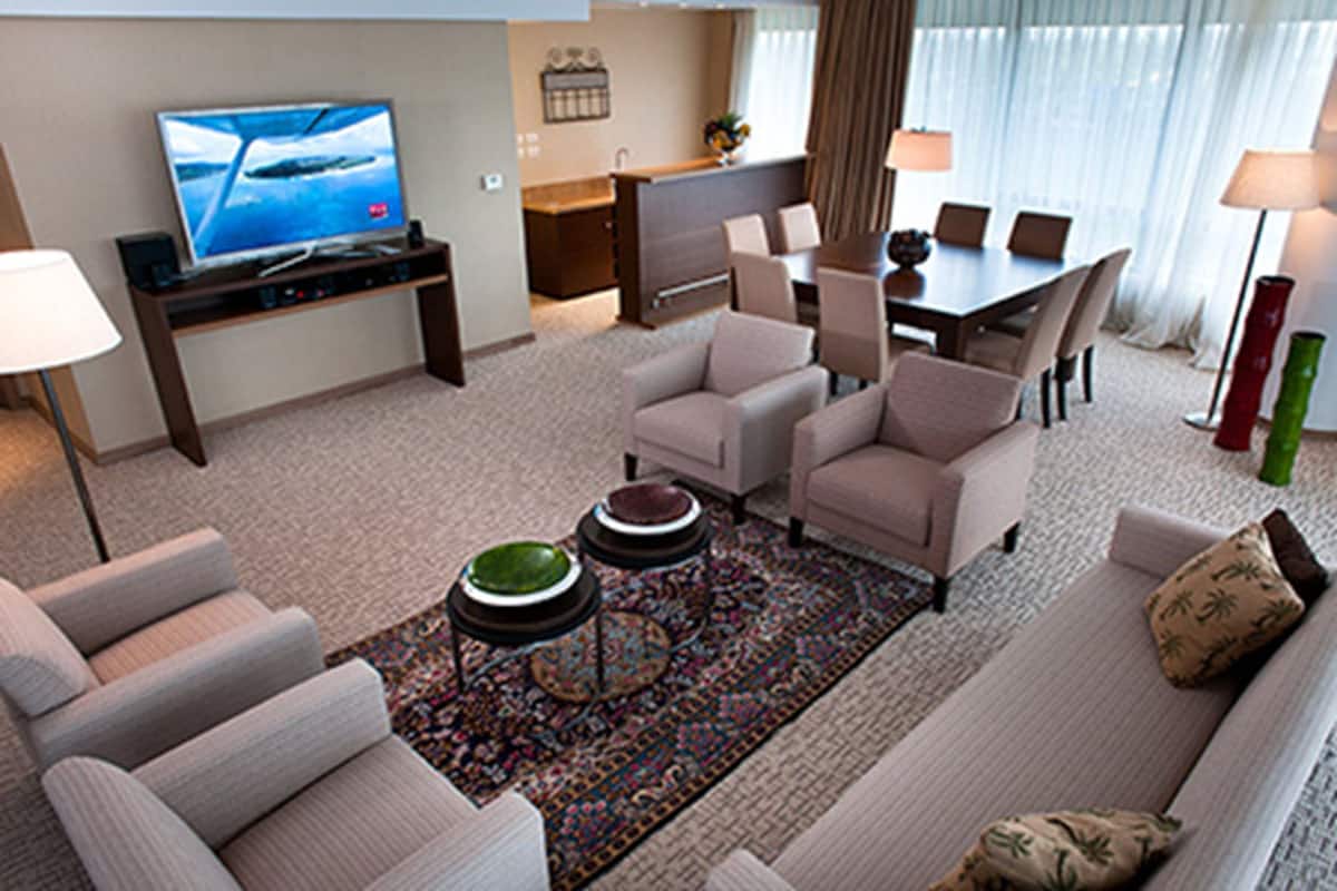 A modern hotel room with a sofa, armchairs, a coffee table, a TV, a dining area, and a kitchen.