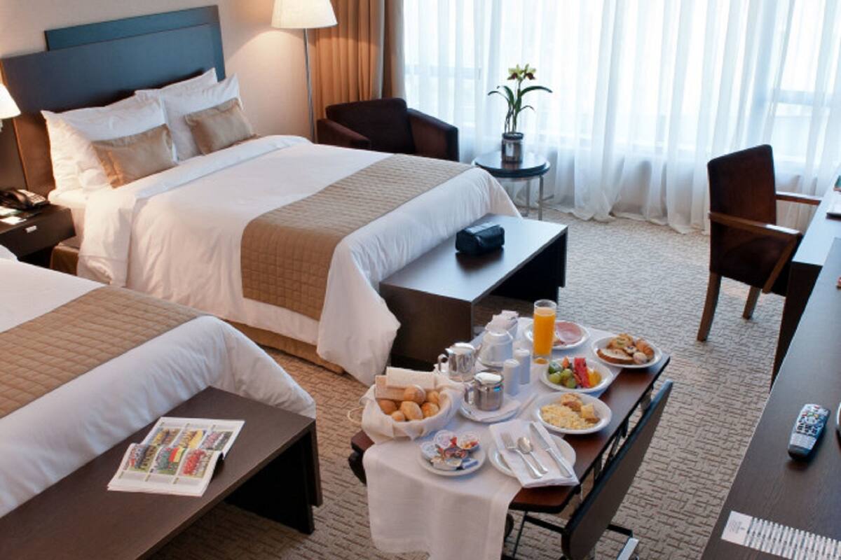 A hotel room with two beds, a dining table with breakfast, a chair, a lamp, and a window with curtains.