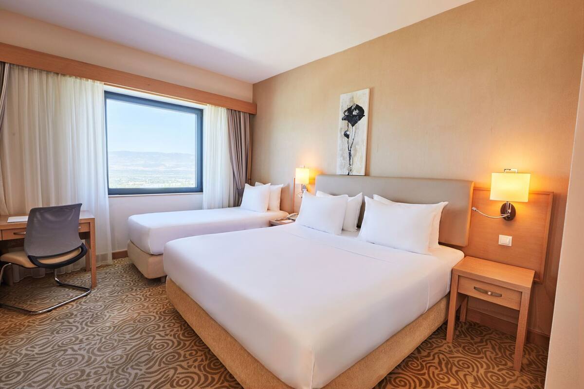 A hotel room with two beds, a desk, a chair, a nightstand, and a wall-mounted lamp.