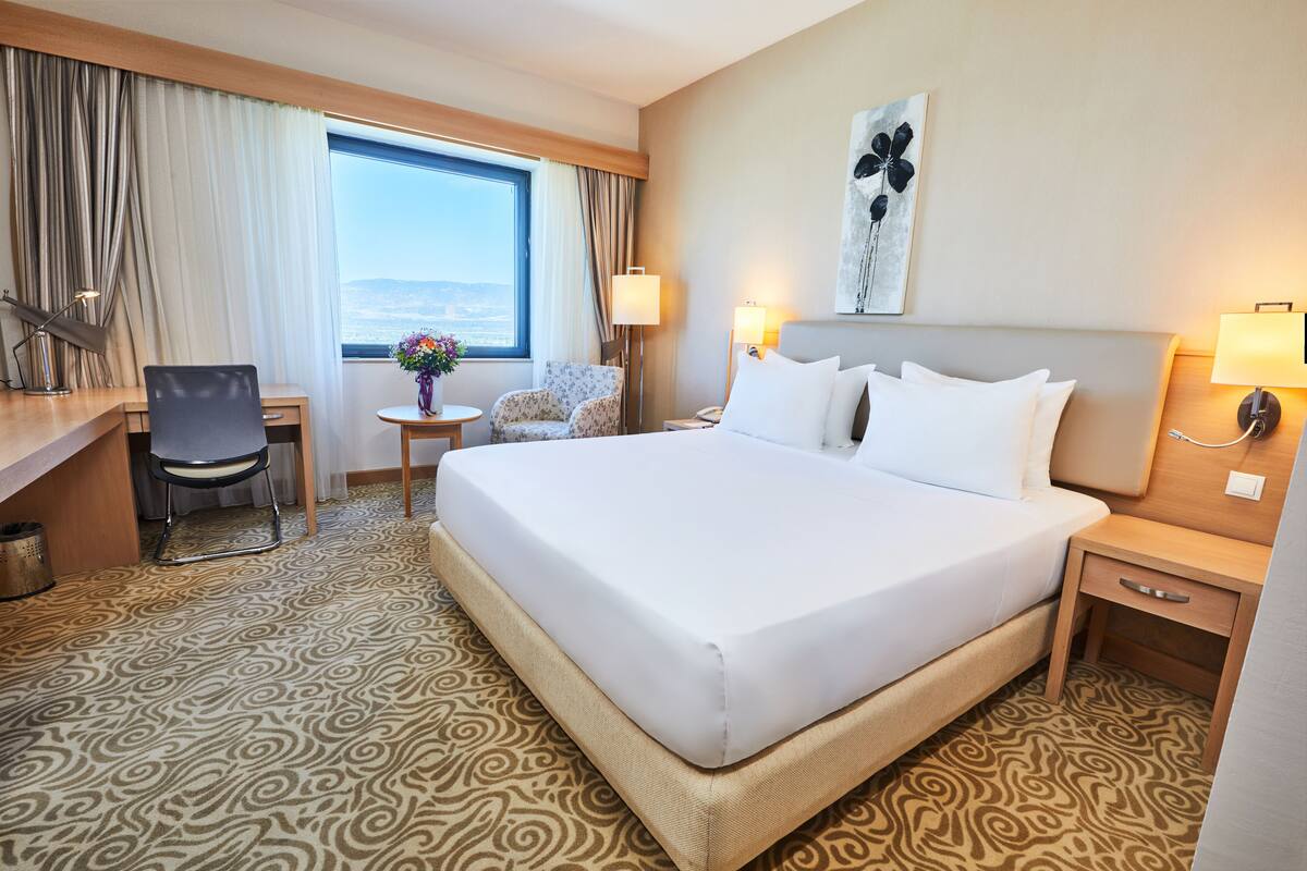 A hotel room with a large bed, a desk, a chair, a small table with a vase, and a view of the outdoors through a large window.