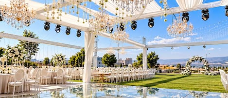 An outdoor event space with a glass floor, white tables, and chandeliers.