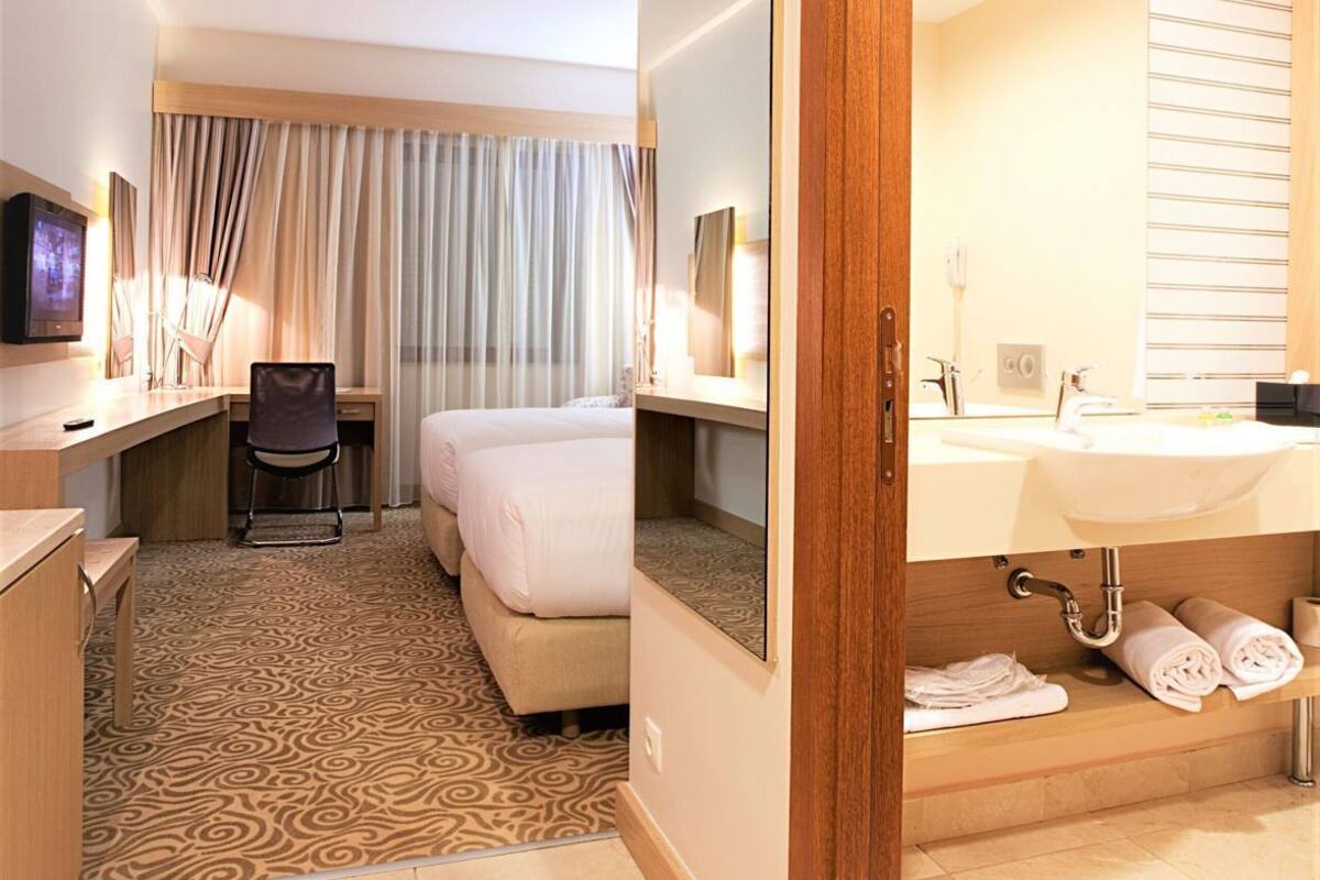 A hotel room with a bed, desk, chair, and a bathroom with a sink and mirror.