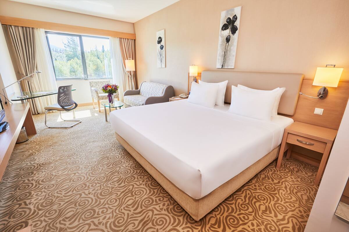 A hotel room with a large bed, a desk with a chair, a small table with a vase of flowers, and a window with curtains.