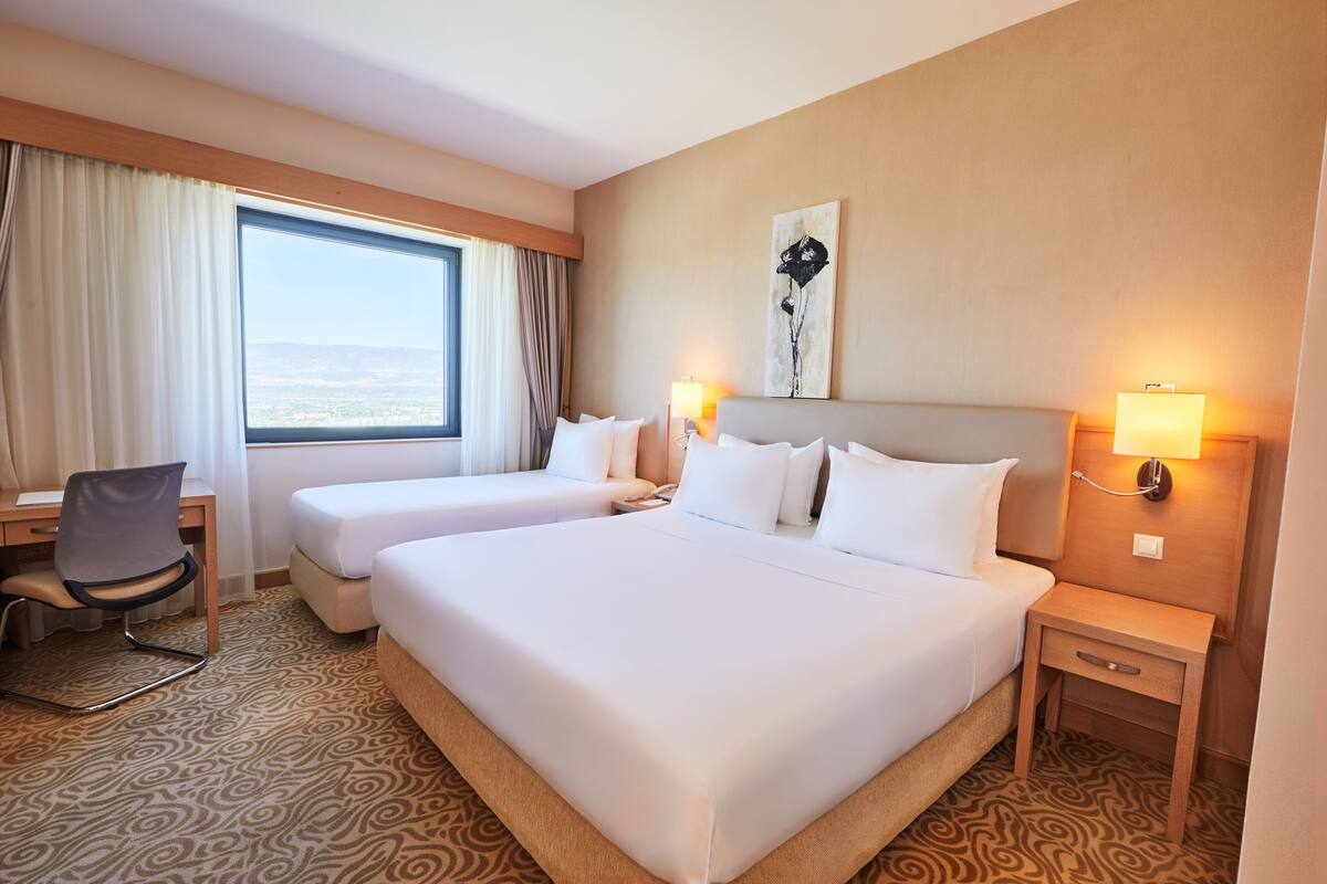 A hotel room with two beds, a desk, a chair, and a large window with curtains.