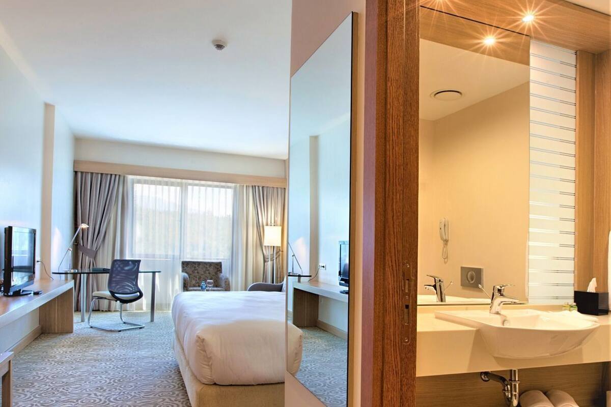A hotel room with a bed, desk, chair, and a bathroom with a sink and mirror.
