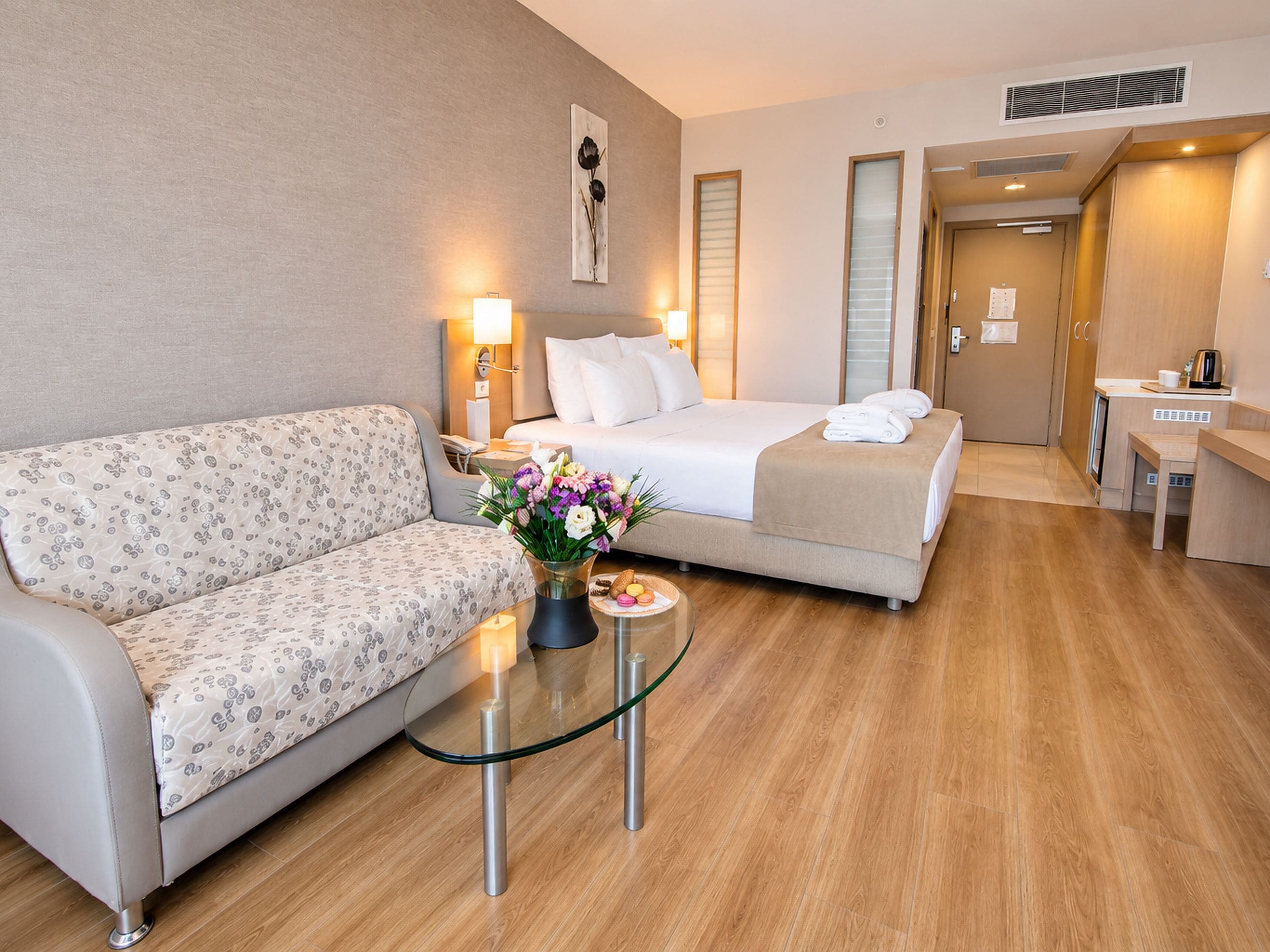 A hotel room with a bed, a sofa, a glass coffee table with flowers, and a kitchenette.