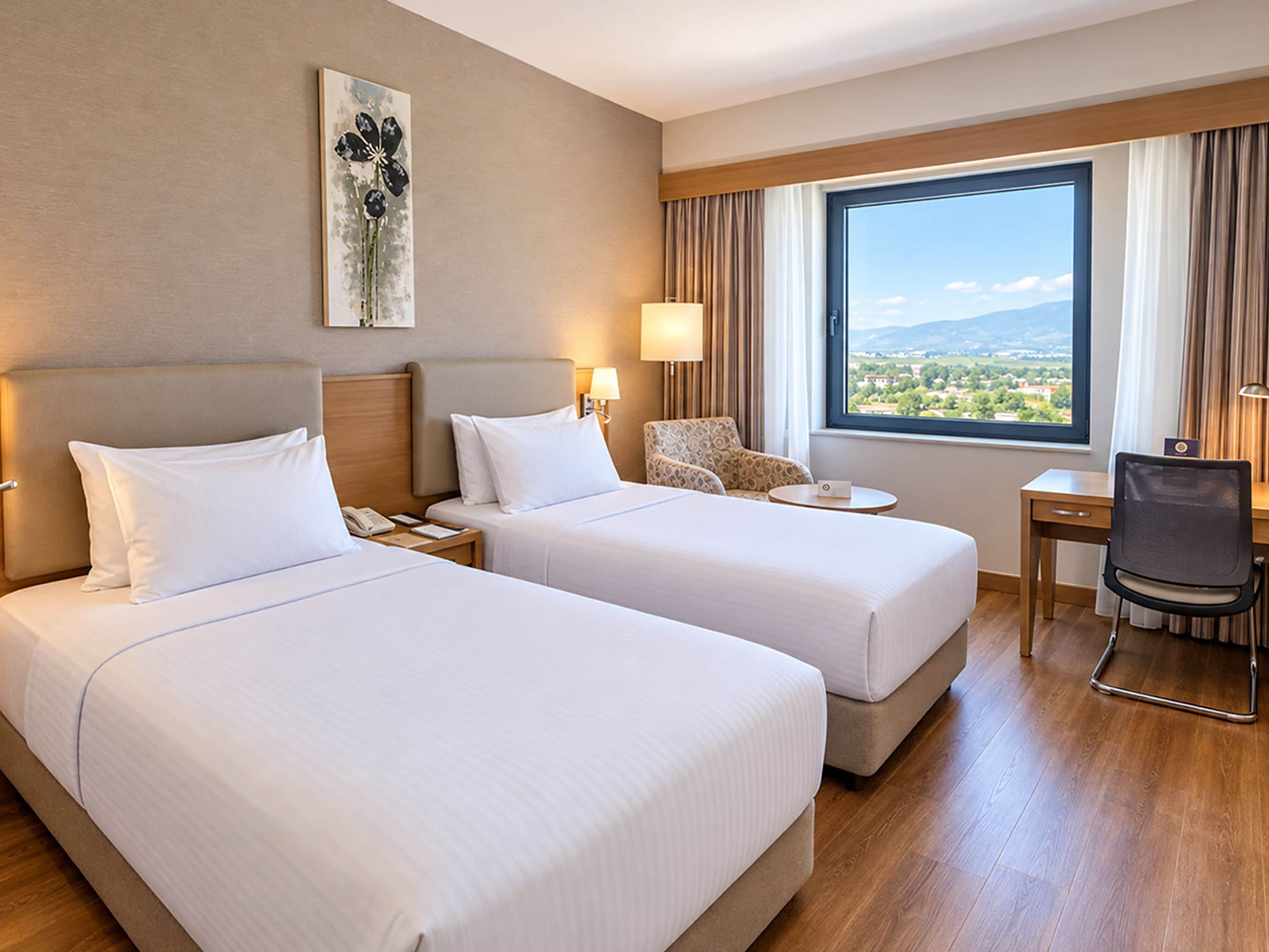 A hotel room with two beds, a desk, a chair, a lamp, and a window with a view.