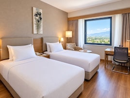 Habitación de hotel con dos camas, un escritorio, una silla, una lámpara y ventana con vistas.