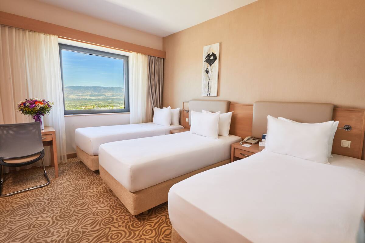 A hotel room with three beds, a desk, a chair, and a large window with a view of the mountains.