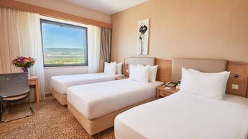 Habitación de hotel con tres camas, un escritorio, una silla y un ventanal con vistas a las montañas.