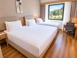 Ein Hotelzimmer mit zwei Betten, einem Schreibtisch und Blick auf einen Wald durch das Fenster.
