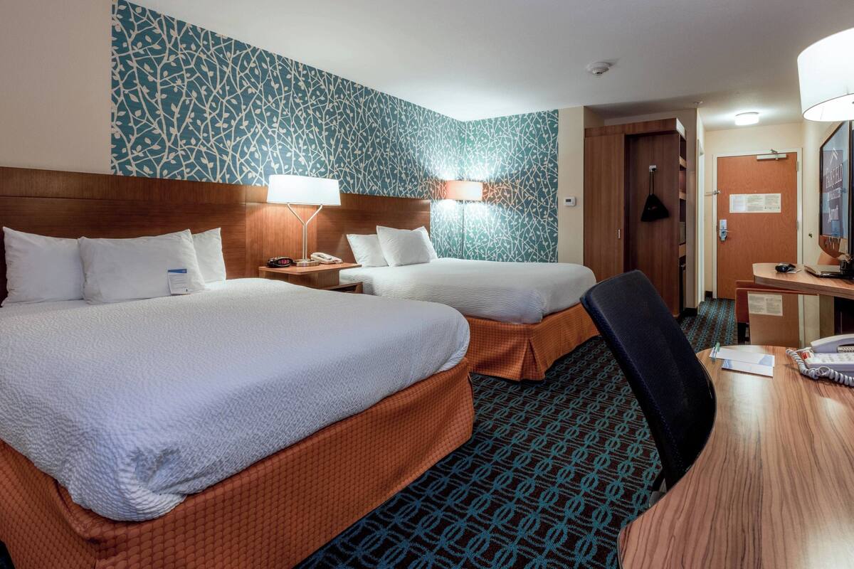 A hotel room with two beds, a desk, and a chair. There are lamps on the desk and a mirror on the wall.