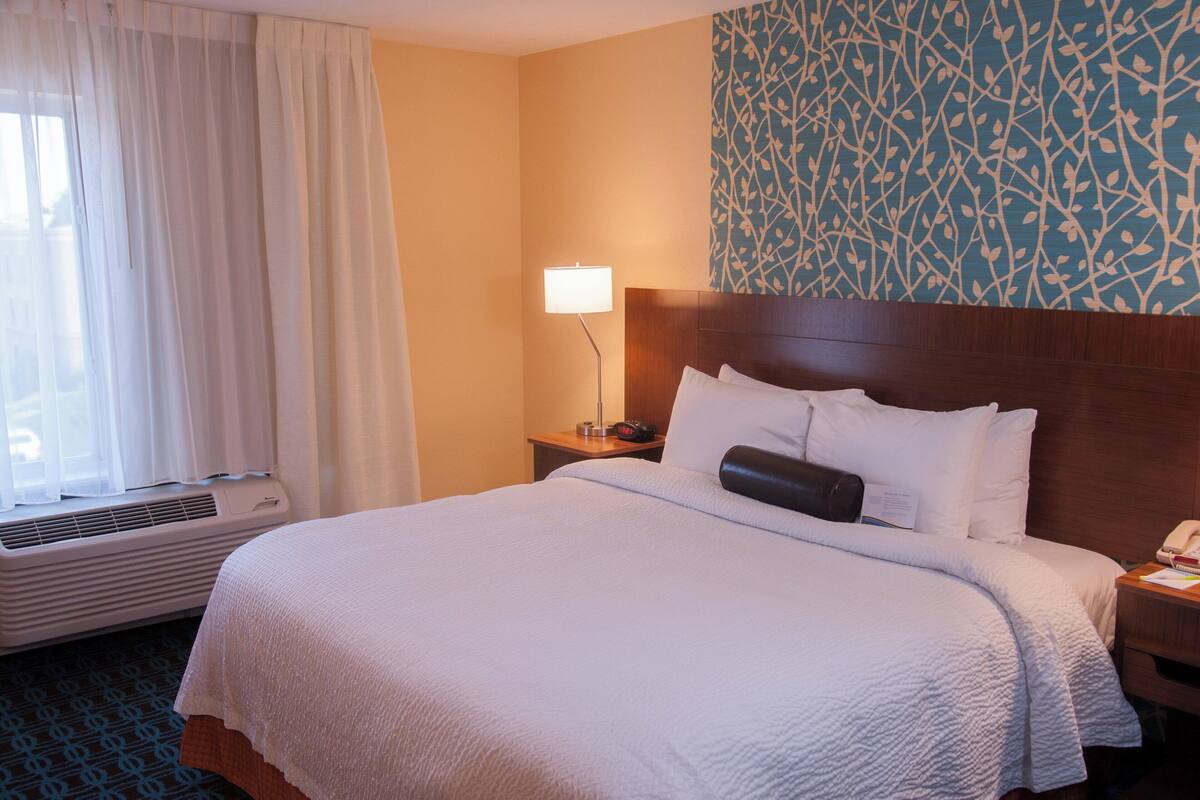 A hotel room with a large bed, a bedside table with a lamp, a wall with a leaf pattern, and a window with curtains.