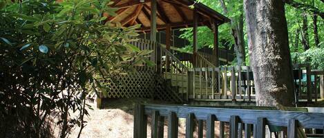 A wooden pavilion with a covered seating area, surrounded by trees and a wooden fence.