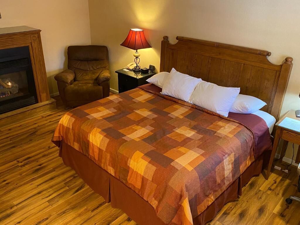 A neatly made bed with a checkered bedspread, a wooden headboard, a leather armchair, a nightstand with a lamp, and a fireplace in the background.