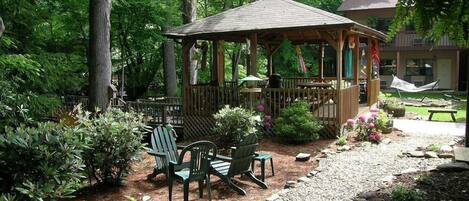 A gazebo with a wooden roof and railings, surrounded by greenery and a gravel path.