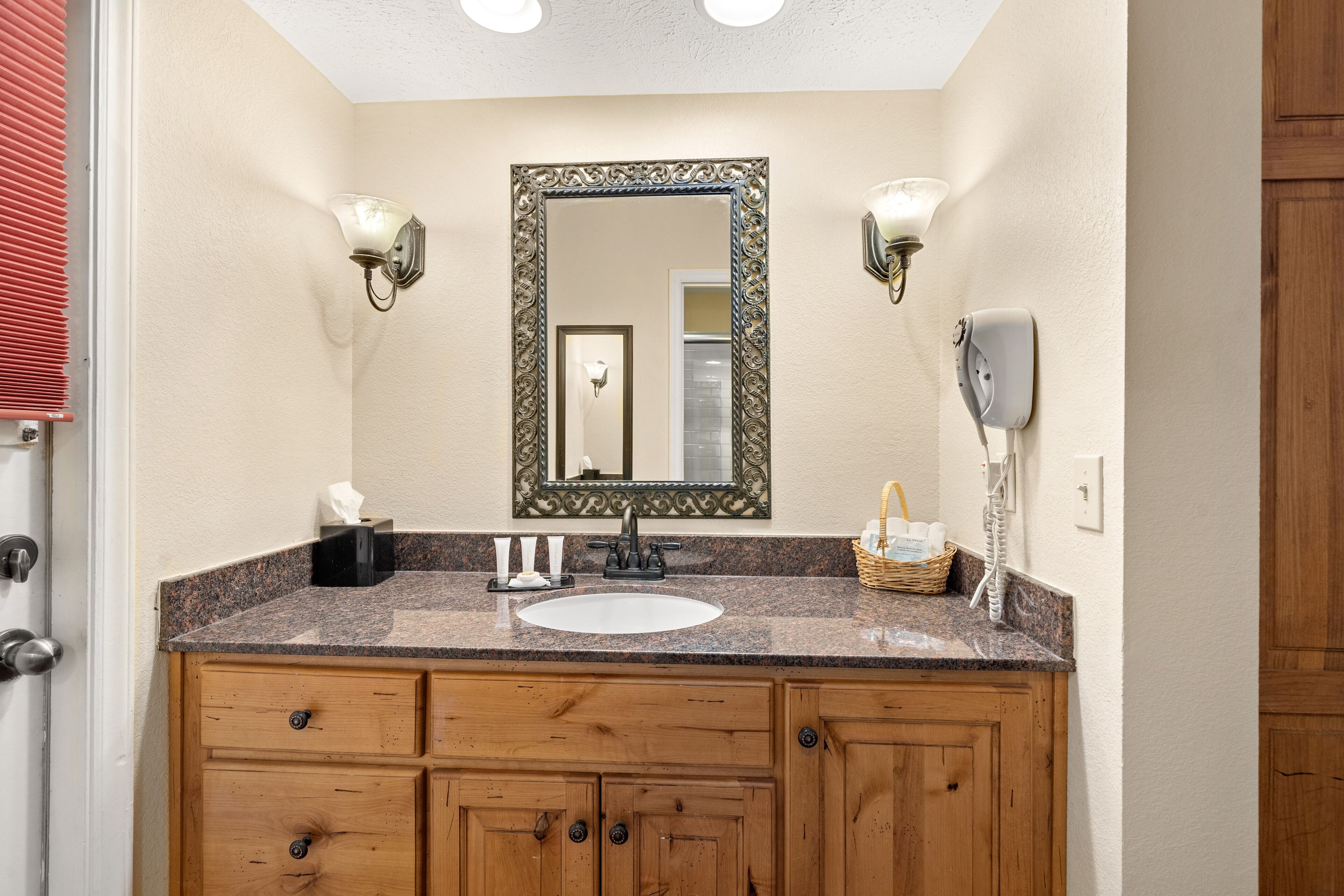A bathroom with a granite countertop, a white sink, wooden cabinets, a large mirror with an ornate frame, two wall-mounted lights, and a hairdryer on the wall.