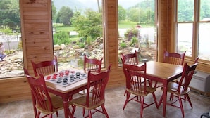 A wooden interior with a table set for two and six red chairs. Large windows offer a view of a garden and a pond.