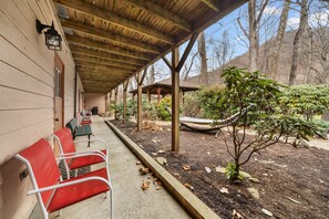 Um espaço coberto ao ar livre com bancos de madeira, uma rede e um pequeno jardim com plantas e árvores.