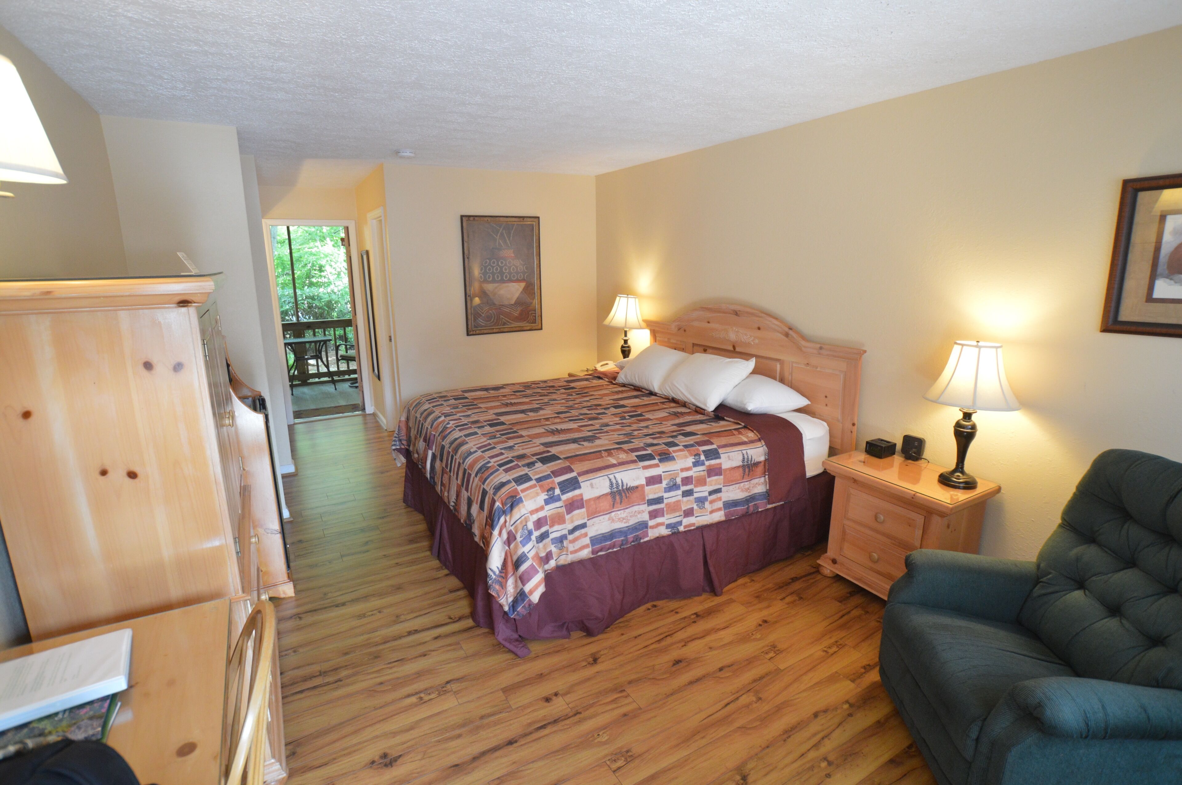 A bedroom with a wooden floor, a bed with a patterned bedspread, a nightstand with a lamp, a chair, and a wooden dresser.