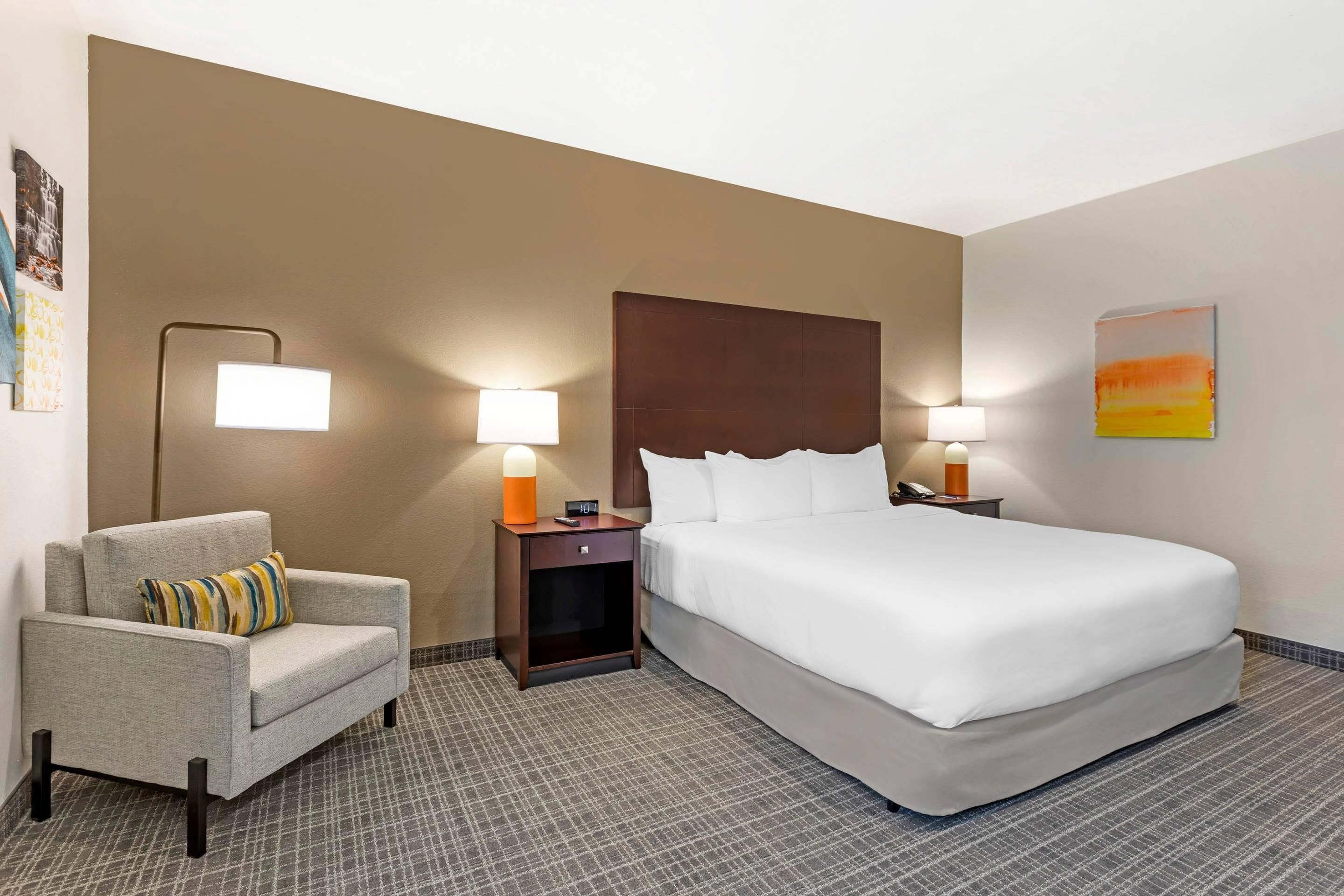 A hotel room with a bed, a chair, two bedside tables with lamps, a wall with a painting, and a patterned carpet.