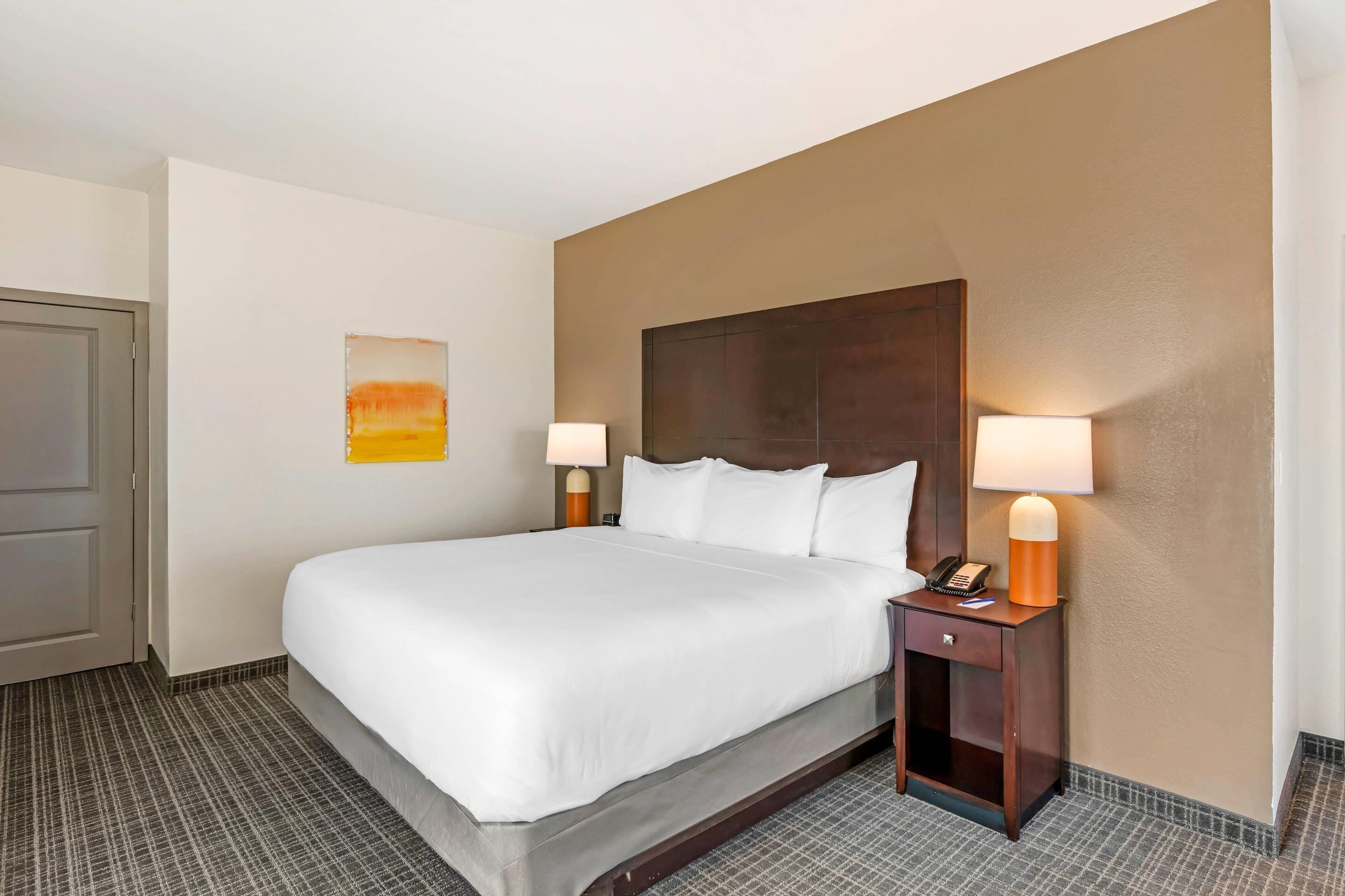 A hotel room with a large bed, two bedside tables with lamps, a door, and a wall-mounted artwork.