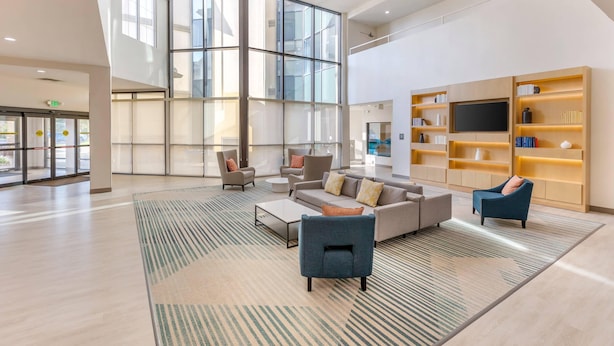 A spacious lobby with a high ceiling, large windows, a seating area with sofas and chairs, a coffee table, and a bookshelf with a flat-screen TV. - Comfort Inn & Suites Verona at Turning Stone Resort Casino (Verona)