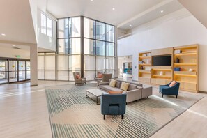 Eine großzügige Lobby mit hoher Decke, großen Fenstern, einer Sitzecke mit Sofas und Sesseln, einem Couchtisch und einem Bücherregal mit Flachbildfernseher.