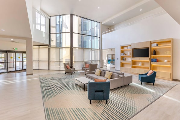 Un vestíbulo amplio con techo alto, ventanas grandes, una zona de estar con sofás y sillones, una mesa de centro y una estantería con televisor de pantalla plana.