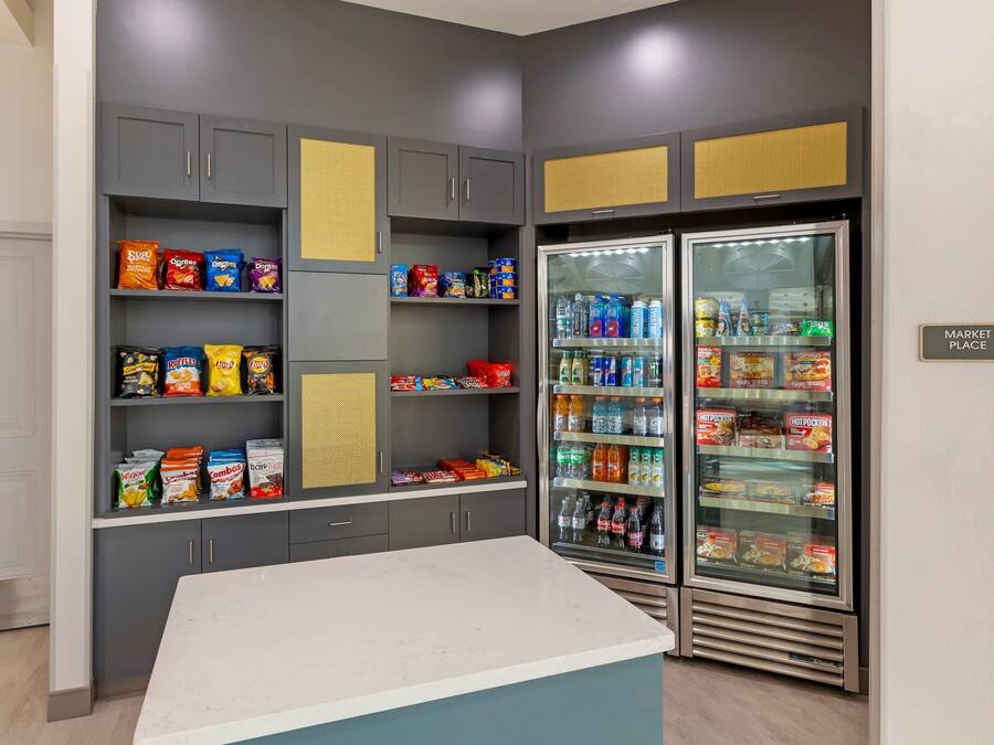 Una kitchenette con un refrigerador lleno de bebidas y aperitivos, una encimera y armarios con diversos alimentos.