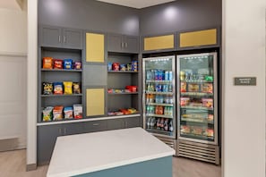 Una kitchenette con un refrigerador lleno de bebidas y aperitivos, una encimera y armarios con diversos alimentos.
