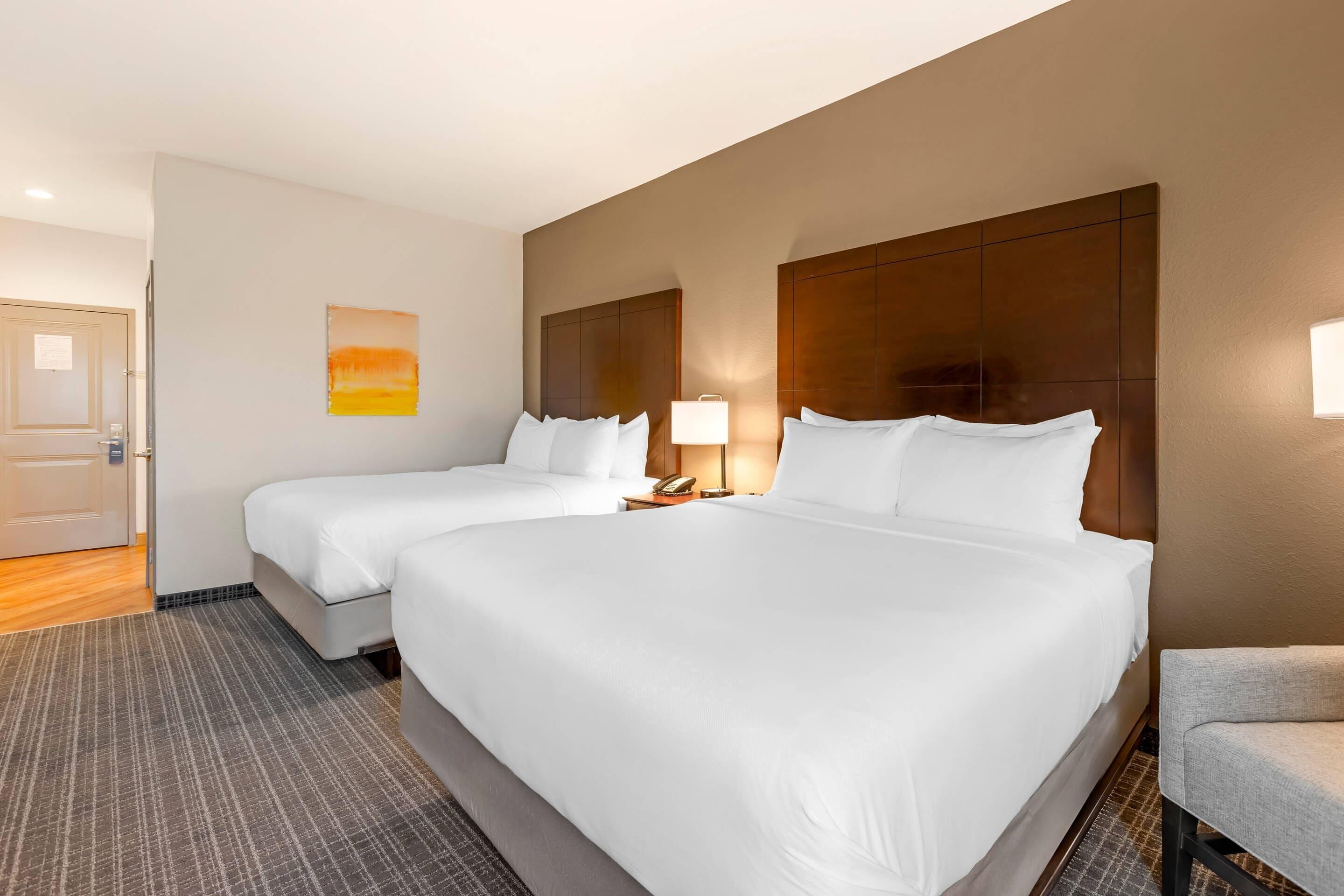 A hotel room with two beds, a nightstand with a lamp, a painting on the wall, and a door.