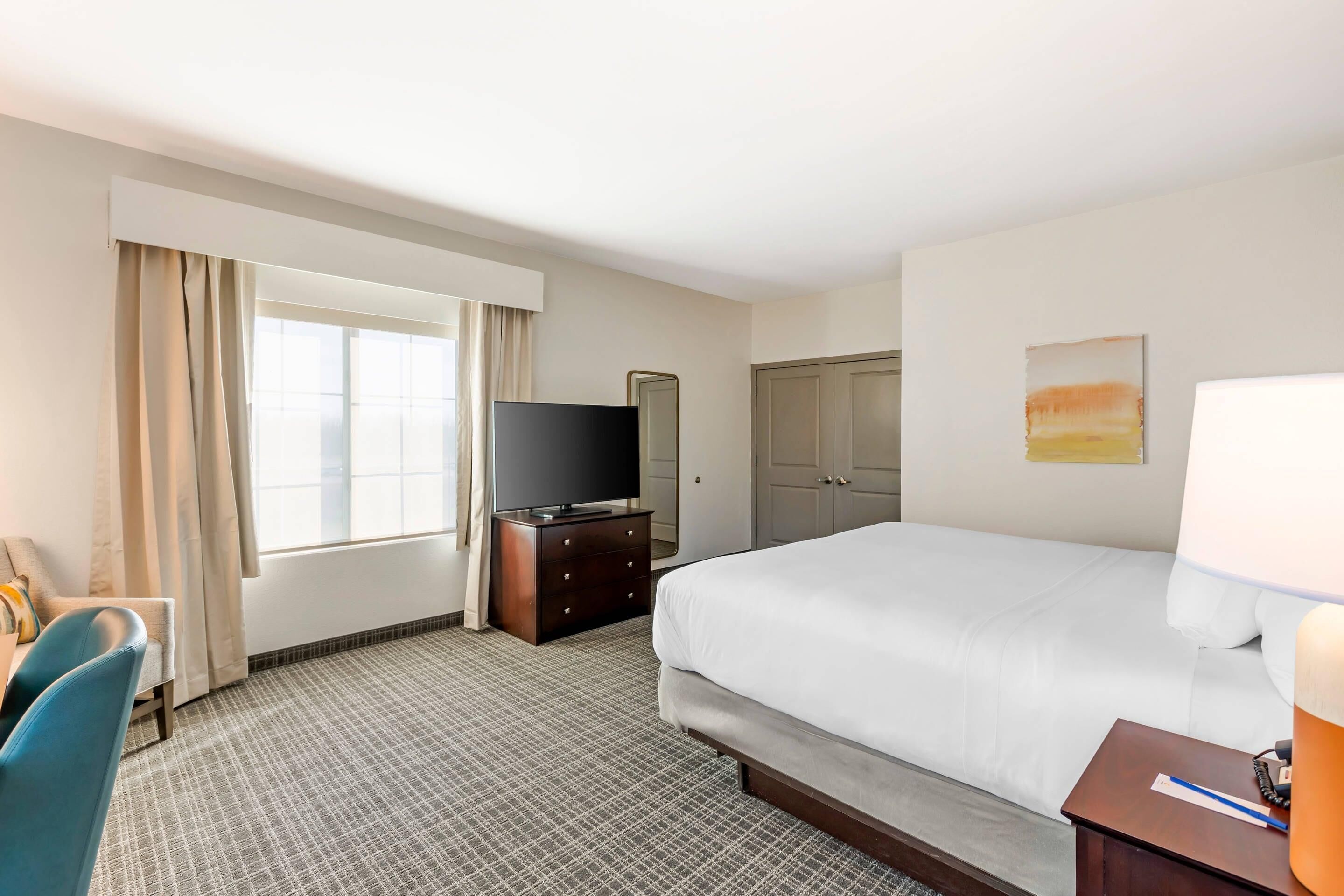 Quarto de hotel com uma cama, uma televisão em uma cômoda, uma escrivaninha com cadeira e uma janela com cortinas.
