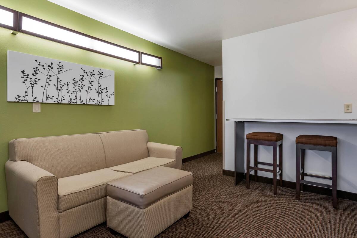 A room with a beige sectional sofa, a wall-mounted artwork, two barstools, and a white counter.