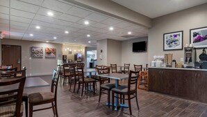 A hotel dining area with multiple tables and chairs, a kitchen area, and a television mounted on the wall.