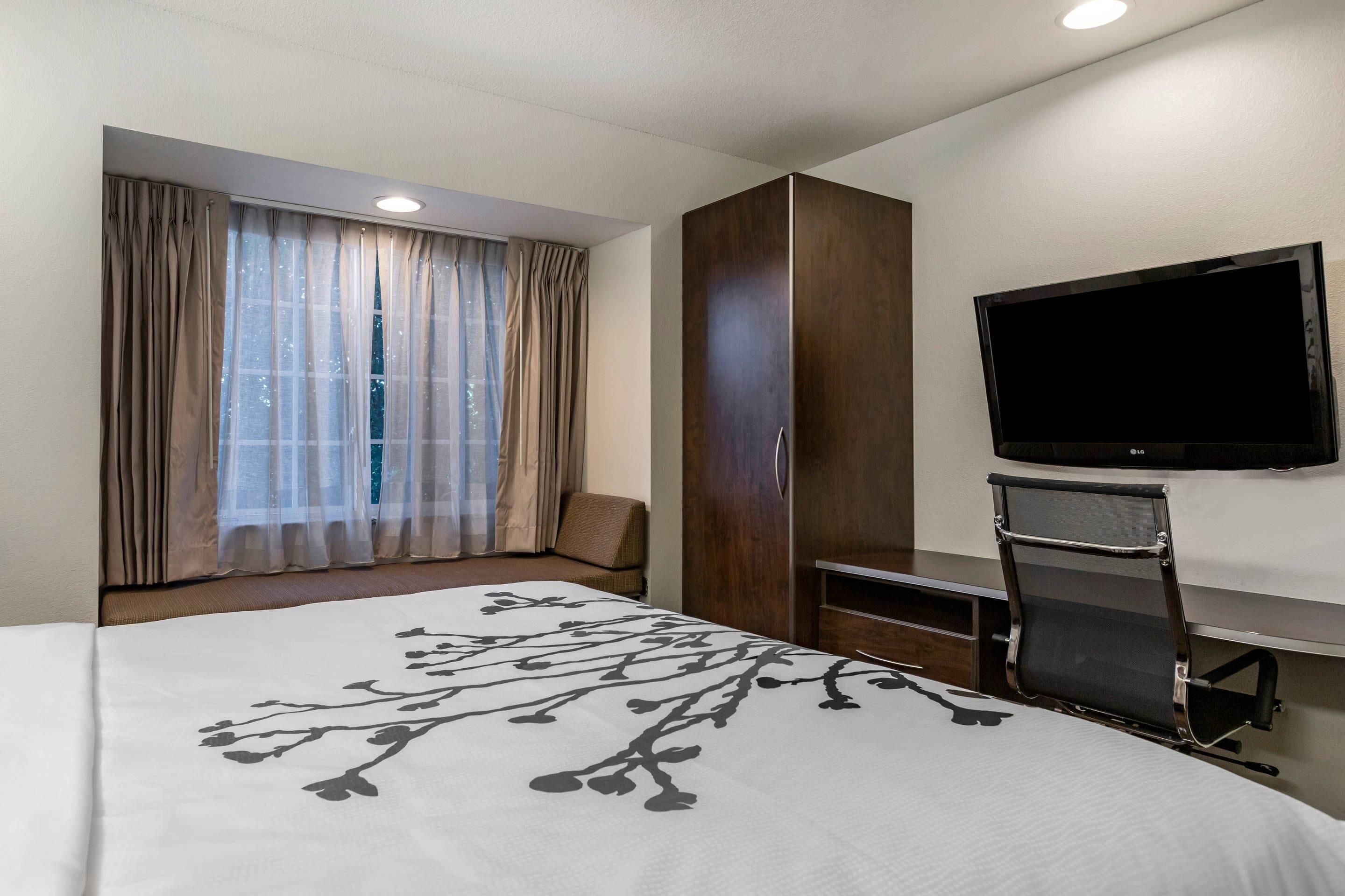 Une chambre d’hôtel avec un lit, un téléviseur fixé au mur, une fenêtre avec des rideaux et une armoire.