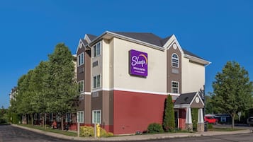 Un hotel Sleep Inn & Suites con un letrero morado llamativo, fachada color beige y roja, y un camino de entrada con un coche rojo estacionado cerca.