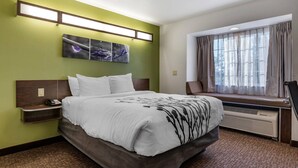 A hotel room with a bed, bedside table, artwork, window with curtains, and an air conditioning unit.