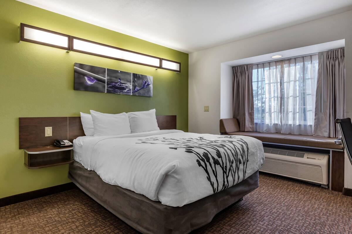 A hotel room with a bed, bedside table, artwork, window with curtains, and an air conditioning unit.