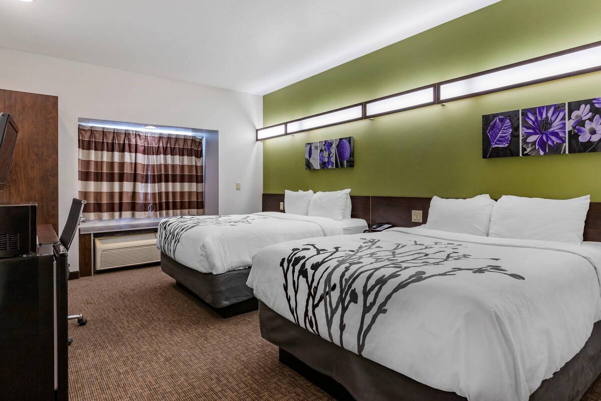 A hotel room with two beds, a flat-screen TV, a window with curtains, and artwork on the wall.