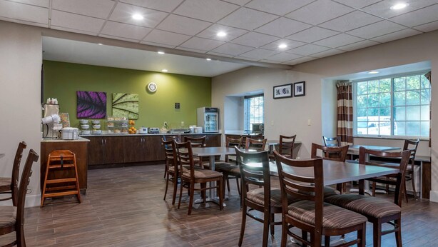 A dining area with wooden chairs and tables, a counter with food items, and a green wall with artwork. - Sleep Inn & Suites Kalamazoo Southwest (Kalamazoo)