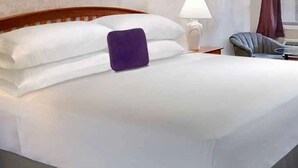 A neatly made bed with white linens and a purple pillow, a wooden headboard, a bedside lamp, and a chair with a round table.