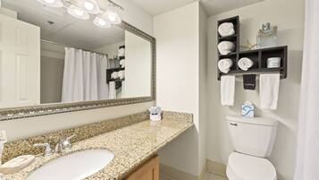 A bathroom with a granite countertop sink, a large mirror, a toilet, and a shelf with towels and toiletries.